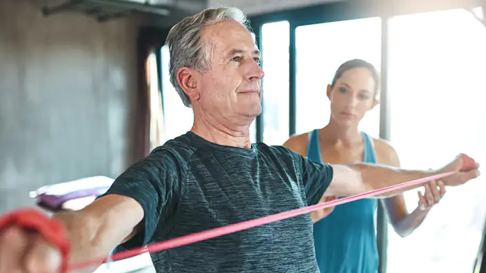 Eine Sport- und Gesundheitstrainerin beobachtet und korrigiert einen älteren Mann bei der Ausführung einer Therabandübung.