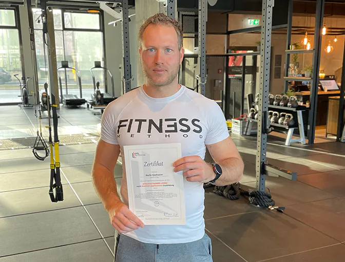 Absolvent Martin zeigt stolz sein Athletiktrainer-Zertifikat in die Kamera, während er im Fitnessstudio steht.