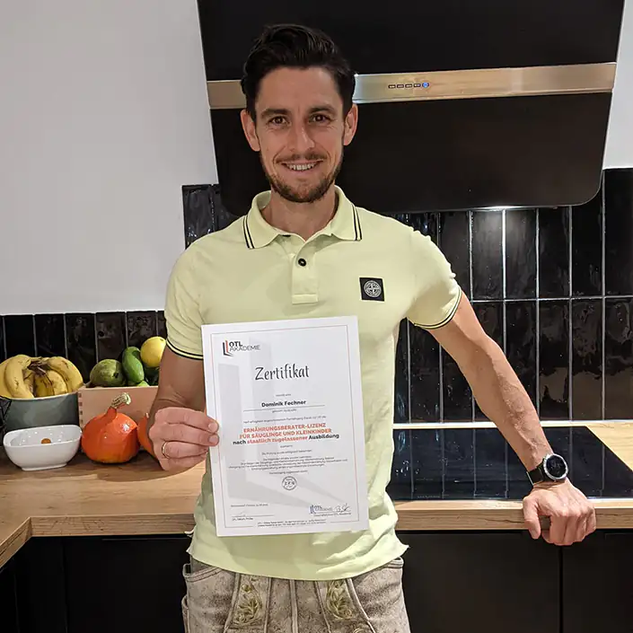 Ernährungsberater für Säuglinge und Kleinkinder Zertifikat (Englisch) Unser Kunde Dominik steht in seiner Küche und hält lächelnd sein Zertifikat des Ernährungsberaters für Säuglinge und Kleinkinder in die Kamera.