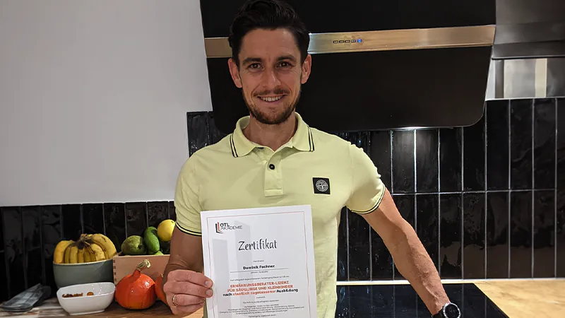 Erfahrung Online Trainer Lizenz Absolvent Dominik steht in einer Küche und zeigt stolz sein Zertifikat zum Ernährungsberater für Säuglinge und Kleinkinder