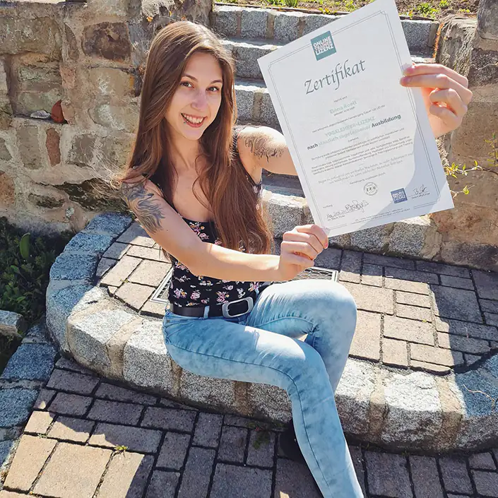 Yogalehrer Zertifikat (Englisch) Unsere Kundin Elena sitzt auf einem Treppenabsatz und zeigt lächelnd ihr Zertifikat nach bestandener Yogalehrer-Ausbildung.